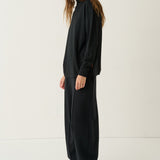 A woman modeling a black oversized long-sleeve top and matching wide-leg pants, paired with white sneakers, standing against a plain white studio background.