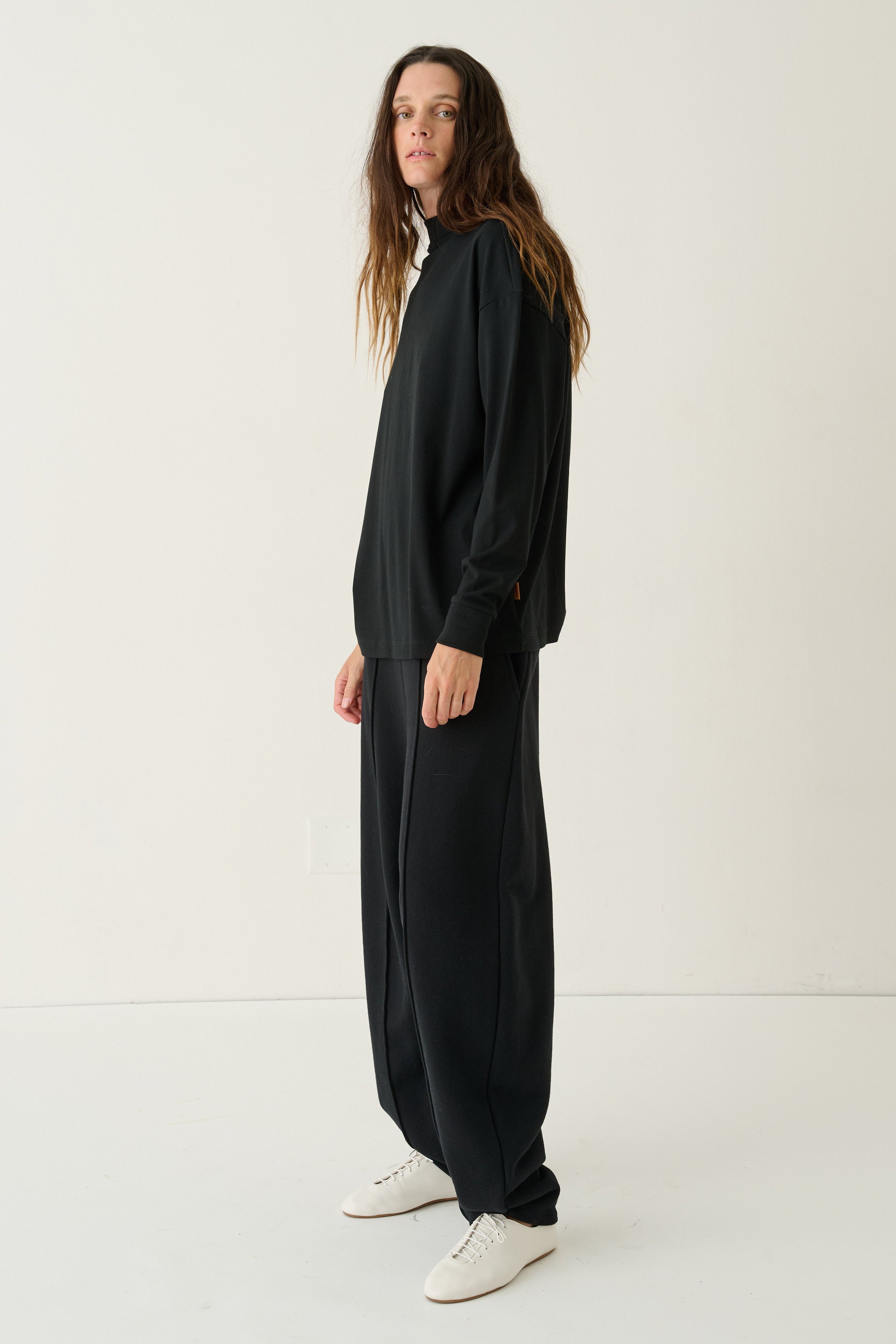 A woman modeling a black oversized long-sleeve top and matching wide-leg pants, paired with white sneakers, standing against a plain white studio background.