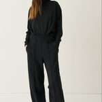 A woman modeling a black long-sleeve top and matching black wide-leg trousers with a tie waist, paired with white sneakers, against a plain white studio background.