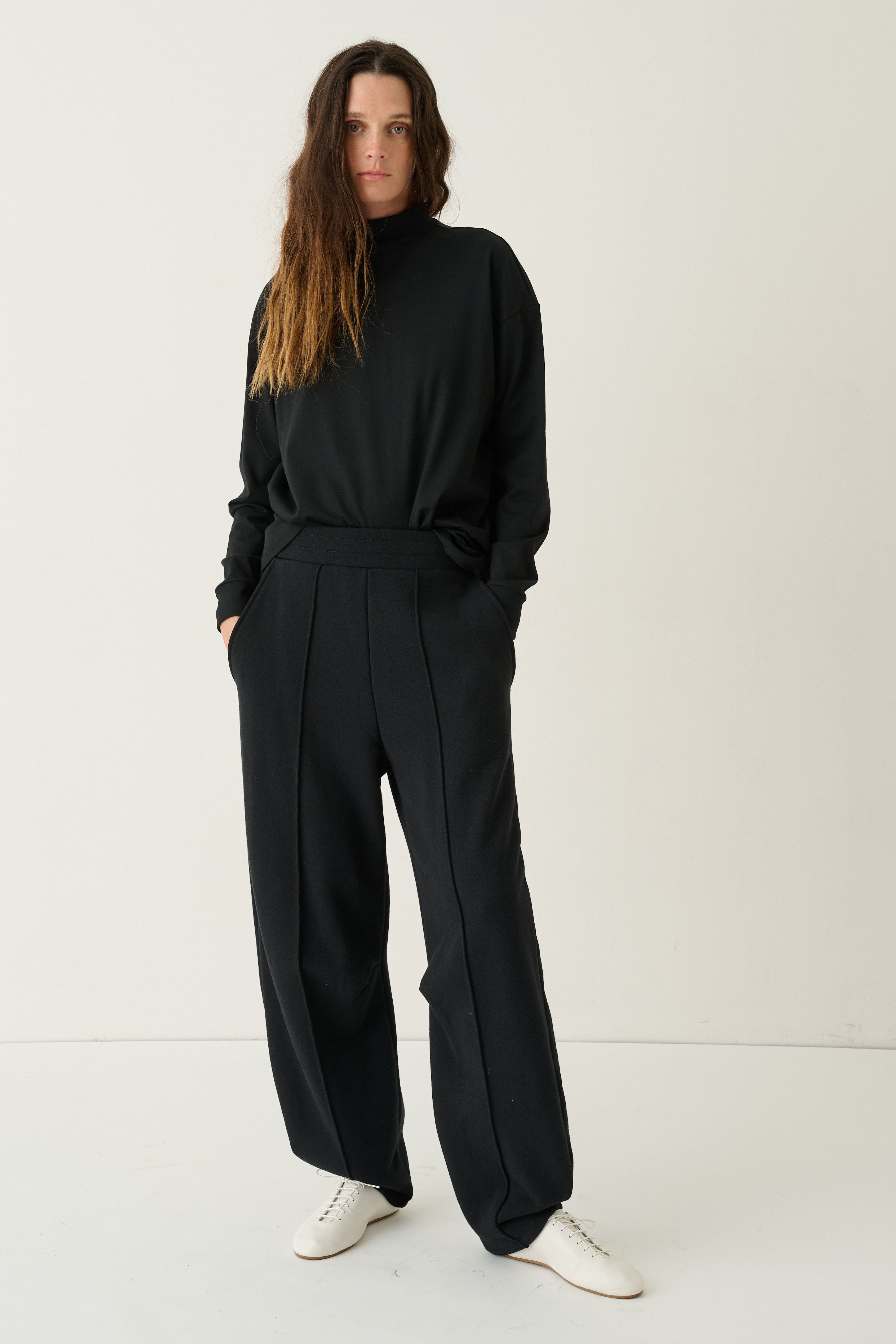 A woman modeling a black long-sleeve top and matching black wide-leg trousers with a tie waist, paired with white sneakers, against a plain white studio background.