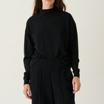 A model wearing a black long-sleeve mock neck top and matching black wide-leg trousers, standing against a plain light gray studio background.