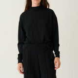 A model wearing a black long-sleeve mock neck top and matching black wide-leg trousers, standing against a plain light gray studio background.