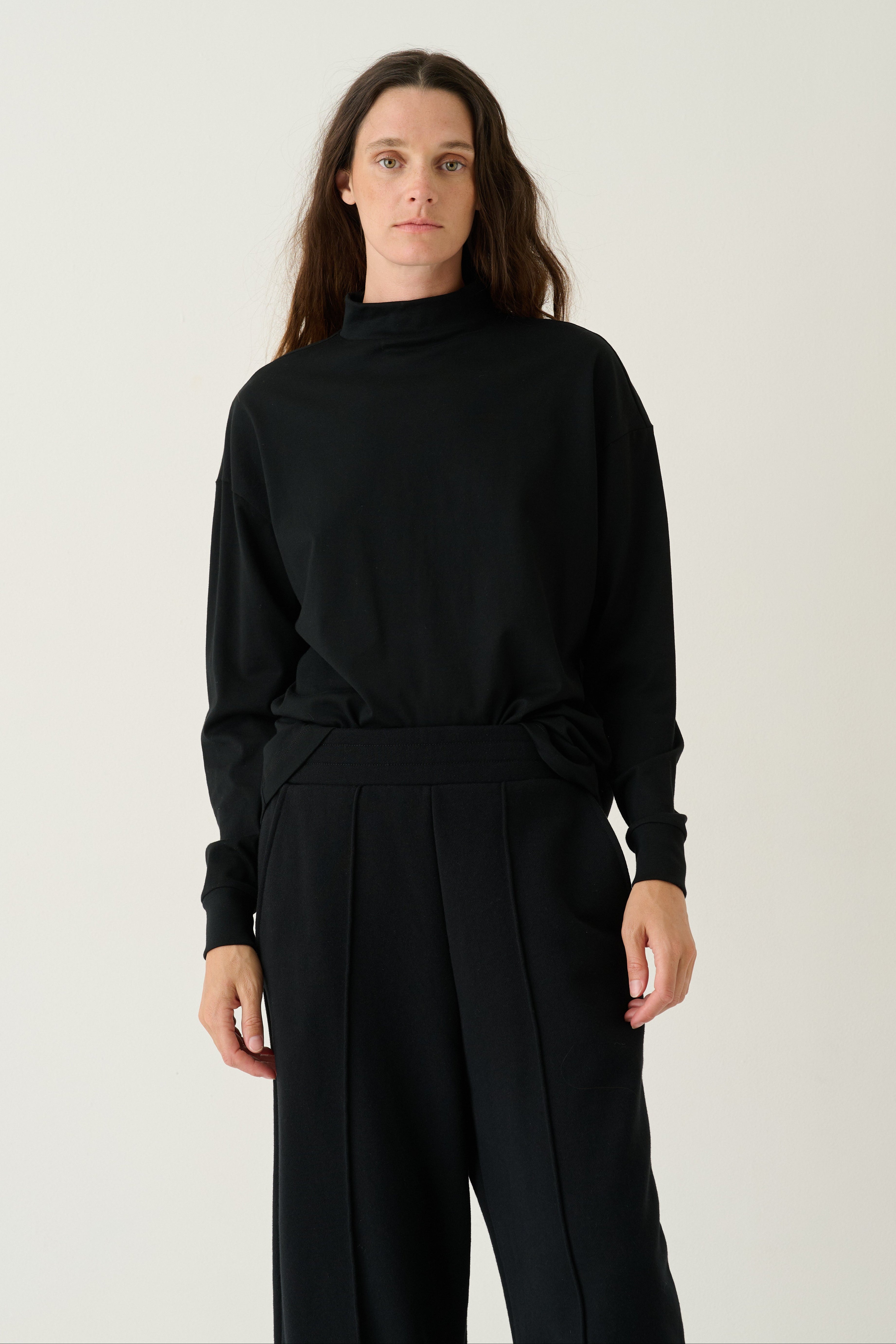 A model wearing a black long-sleeve mock neck top and matching black wide-leg trousers, standing against a plain light gray studio background.