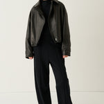 A model stands against a plain light background wearing a dark brown leather jacket over a black top and black wide-leg trousers, paired with white sneakers.