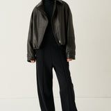 A model stands against a plain light background wearing a dark brown leather jacket over a black top and black wide-leg trousers, paired with white sneakers.