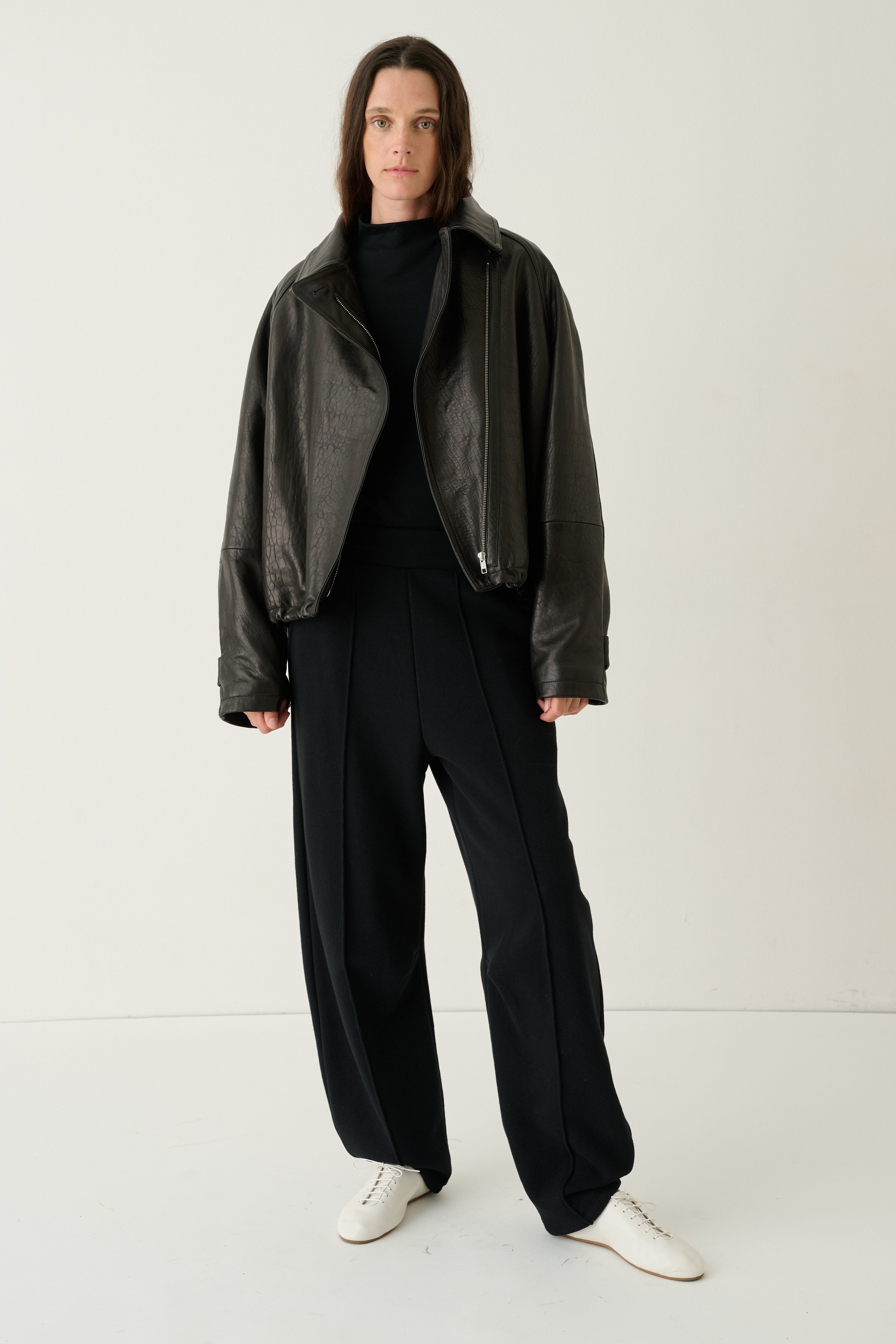 A model stands against a plain light background wearing a dark brown leather jacket over a black top and black wide-leg trousers, paired with white sneakers.
