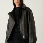 A model wearing a black leather moto jacket with asymmetrical zippers and a high collar, styled over a black top and black trousers, set against a plain light gray studio background.