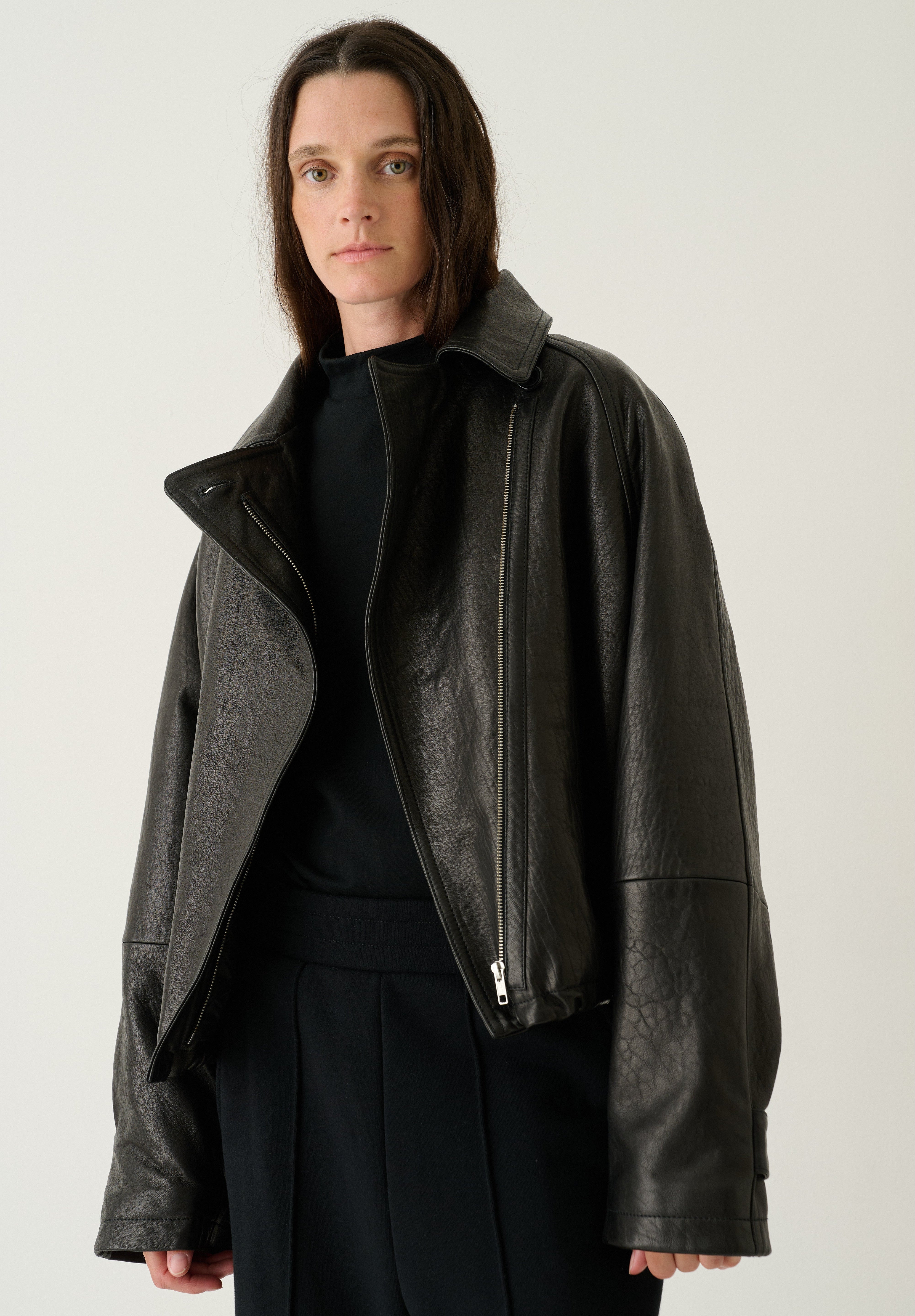 A model wearing a black leather moto jacket with asymmetrical zippers and a high collar, styled over a black top and black trousers, set against a plain light gray studio background.
