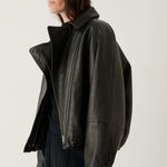 A woman wearing a black leather moto jacket with an asymmetrical zipper and high collar, paired with a black top and dark trousers, photographed against a plain light gray studio background.