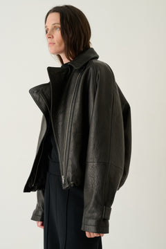 A woman wearing a black leather moto jacket with an asymmetrical zipper and high collar, paired with a black top and dark trousers, photographed against a plain light gray studio background.