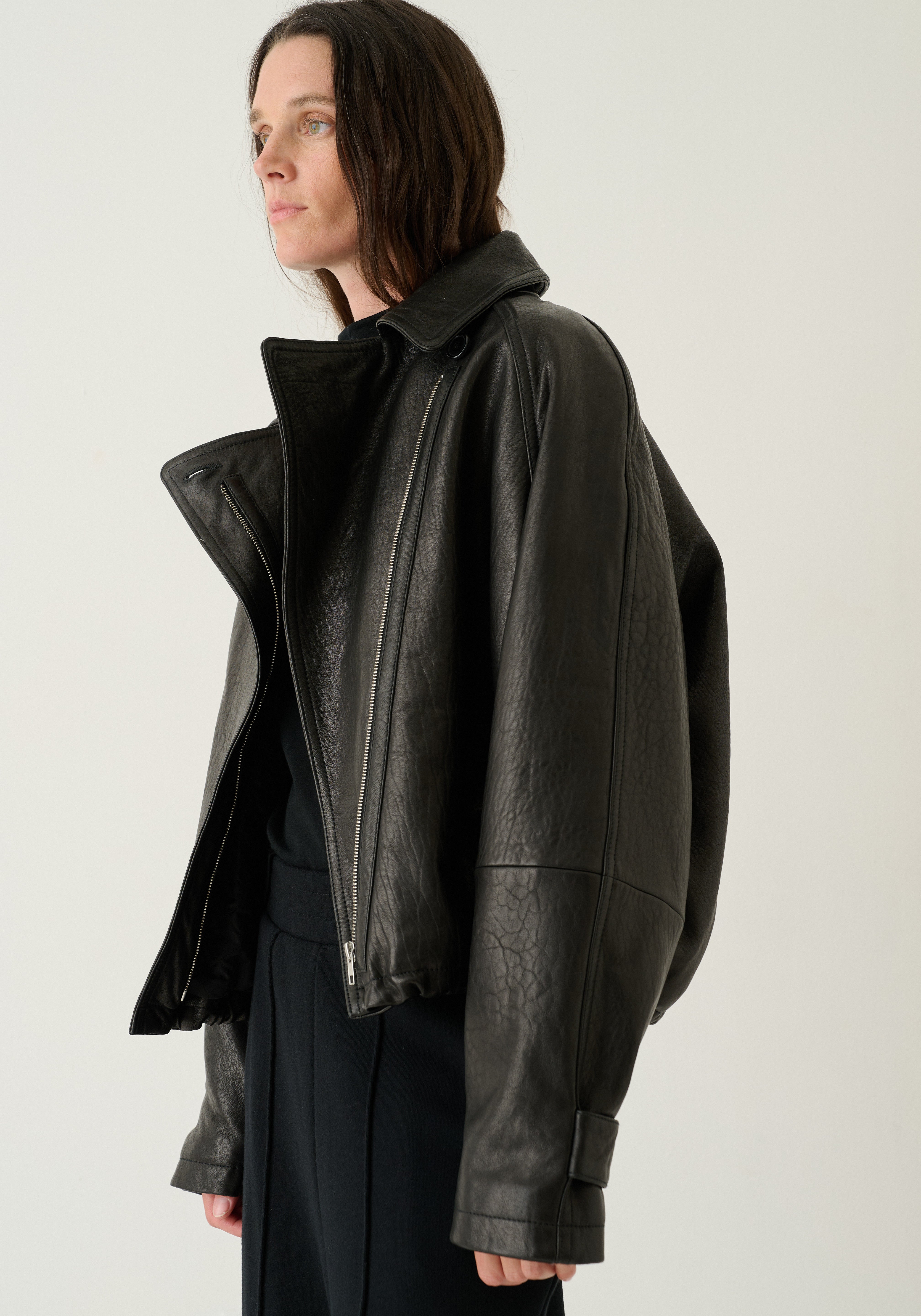 A woman wearing a black leather moto jacket with an asymmetrical zipper and high collar, paired with a black top and dark trousers, photographed against a plain light gray studio background.