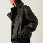 A model wearing a black leather jacket with a high collar and gathered hem, paired with dark trousers, photographed against a plain light gray studio background.