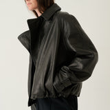 A model wearing a black leather jacket with a high collar and gathered hem, paired with dark trousers, photographed against a plain light gray studio background.