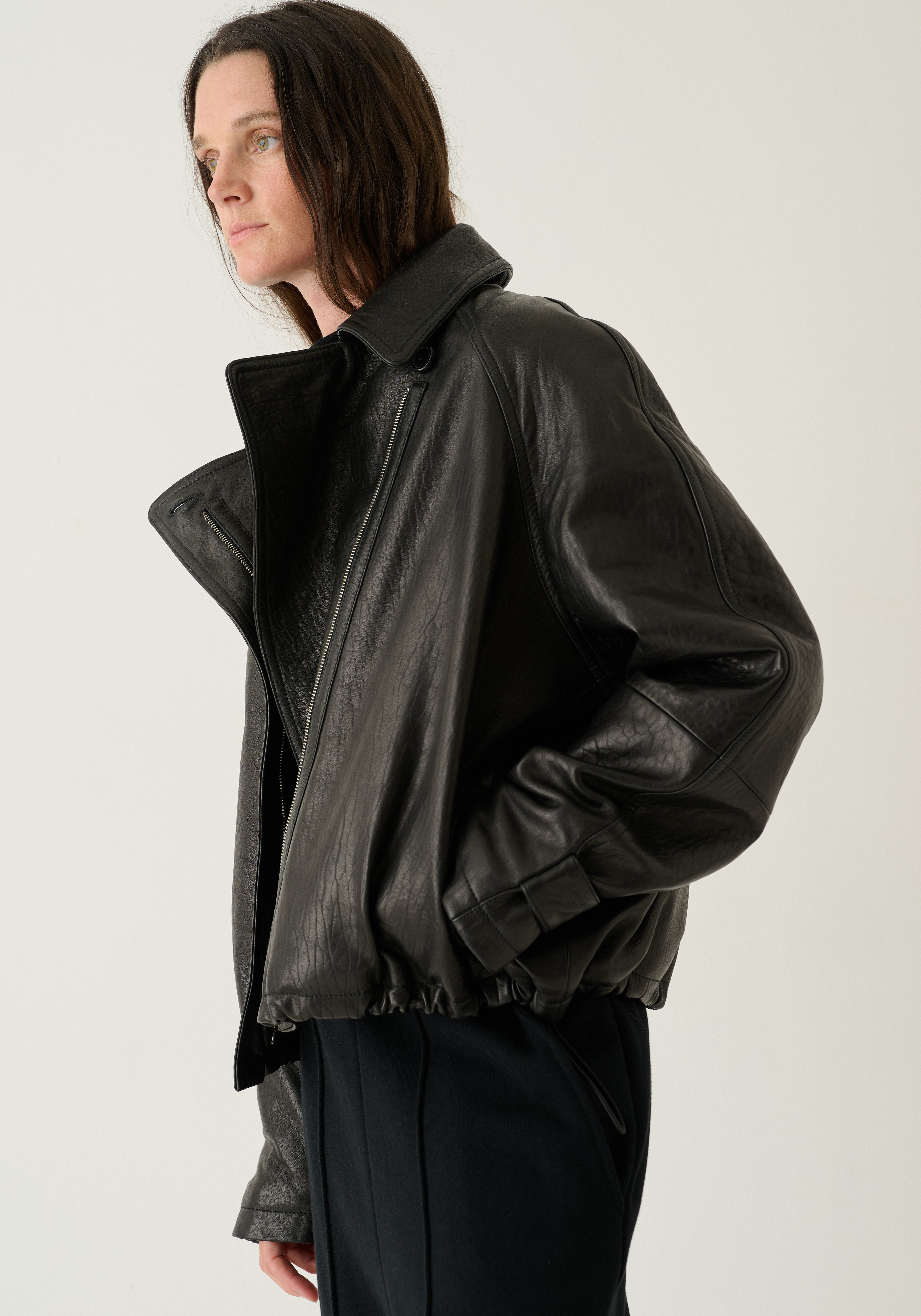 A model wearing a black leather jacket with a high collar and gathered hem, paired with dark trousers, photographed against a plain light gray studio background.