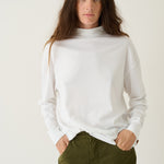 A woman wearing a white long-sleeve mock neck top and olive green pants, standing against a plain off-white studio background.