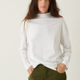 A woman wearing a white long-sleeve mock neck top and olive green pants, standing against a plain off-white studio background.
