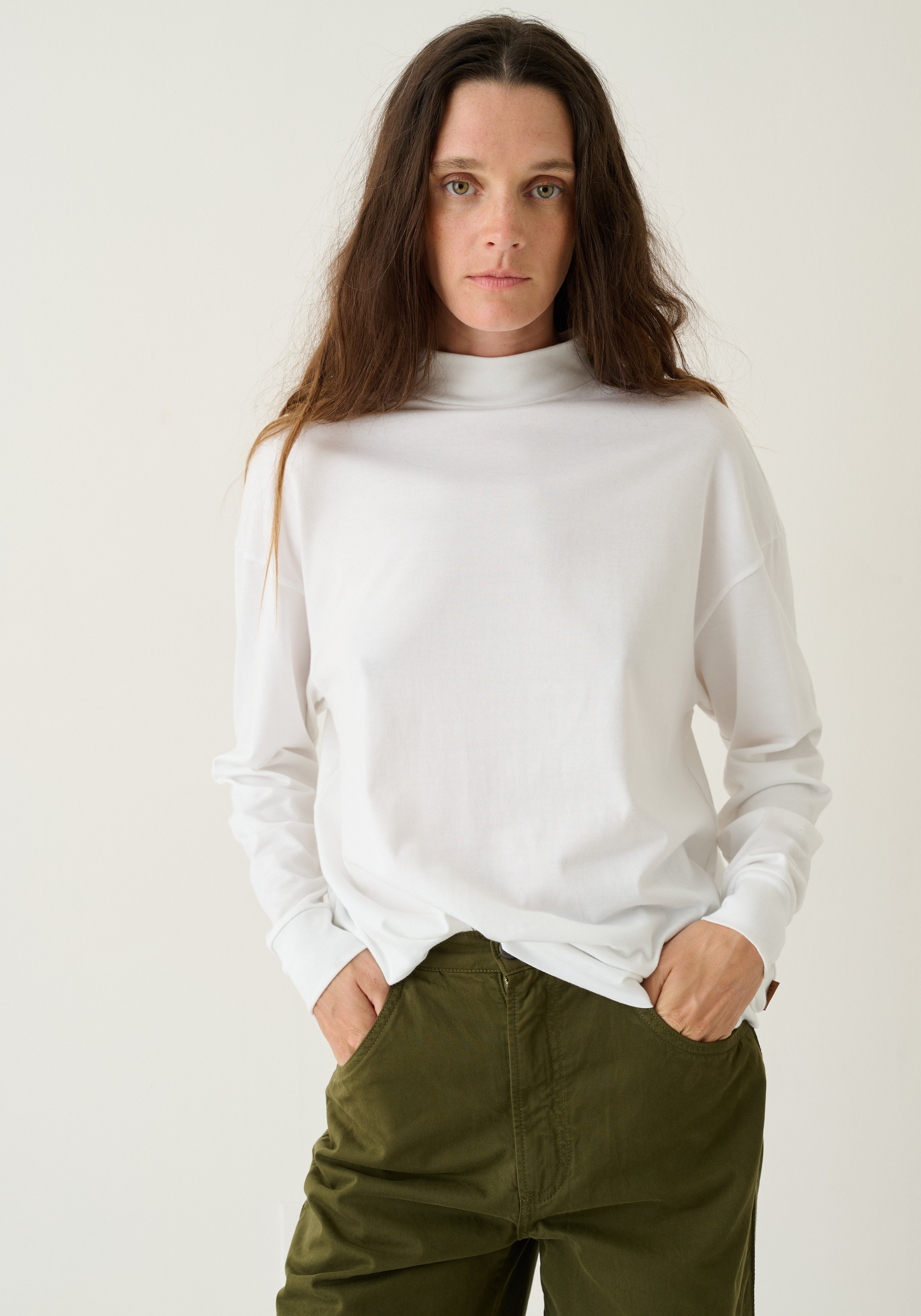 A woman wearing a white long-sleeve mock neck top and olive green pants, standing against a plain off-white studio background.