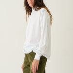 A woman modeling a white long-sleeve top with voluminous balloon sleeves and olive green trousers, standing against a plain off-white studio background.