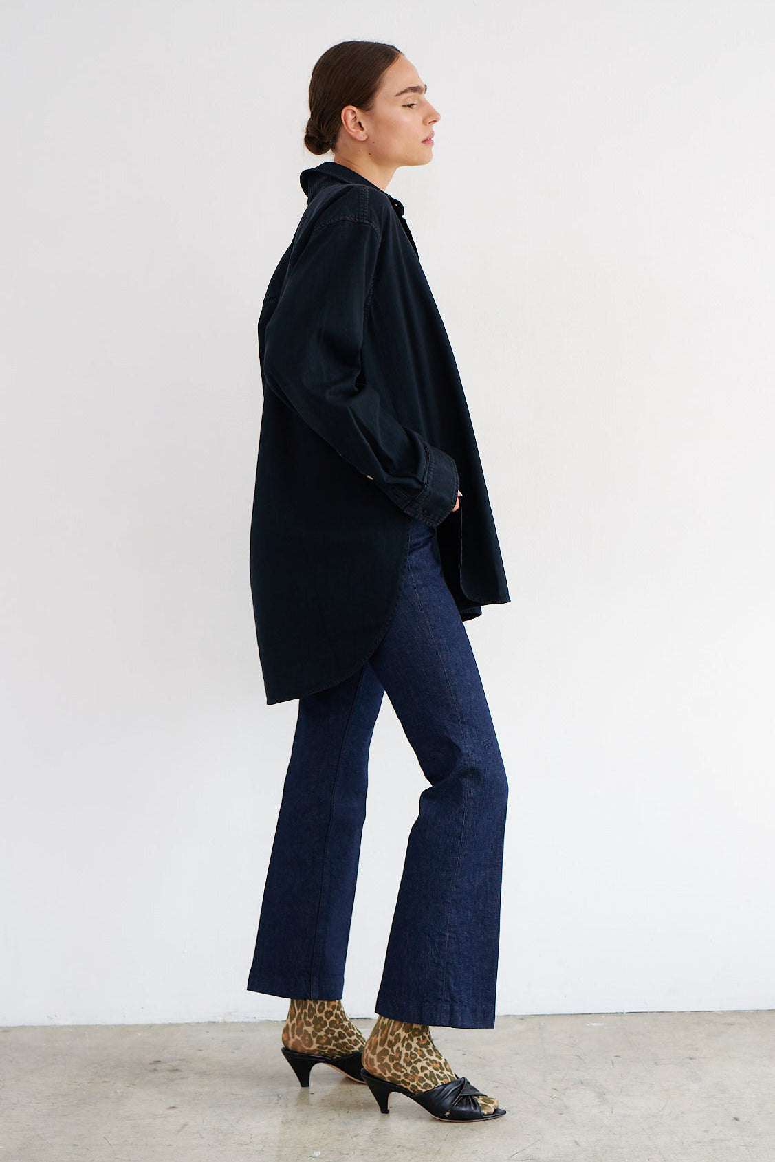A woman stands in profile wearing an oversized dark navy or black longline shirt-jacket, dark blue flared jeans, and leopard-print heeled shoes against a plain light gray wall and concrete floor.