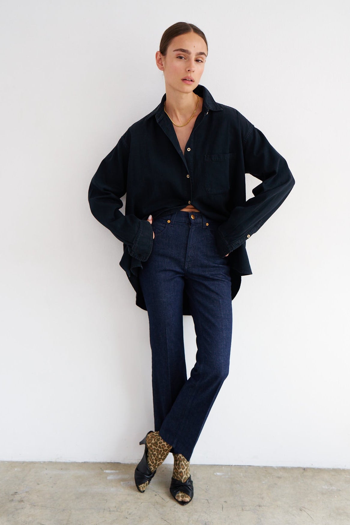 A model stands against a white wall wearing a dark navy blue button-up shirt, matching dark navy trousers, and leopard-print pointed-toe heels.