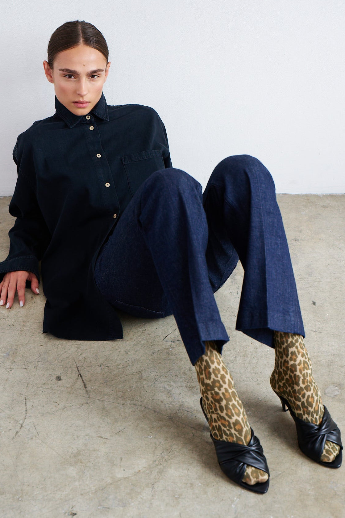 A model sits on a concrete floor against a white wall, wearing a dark navy shirt and matching navy trousers, styled with leopard-print socks and black peep-toe heels.