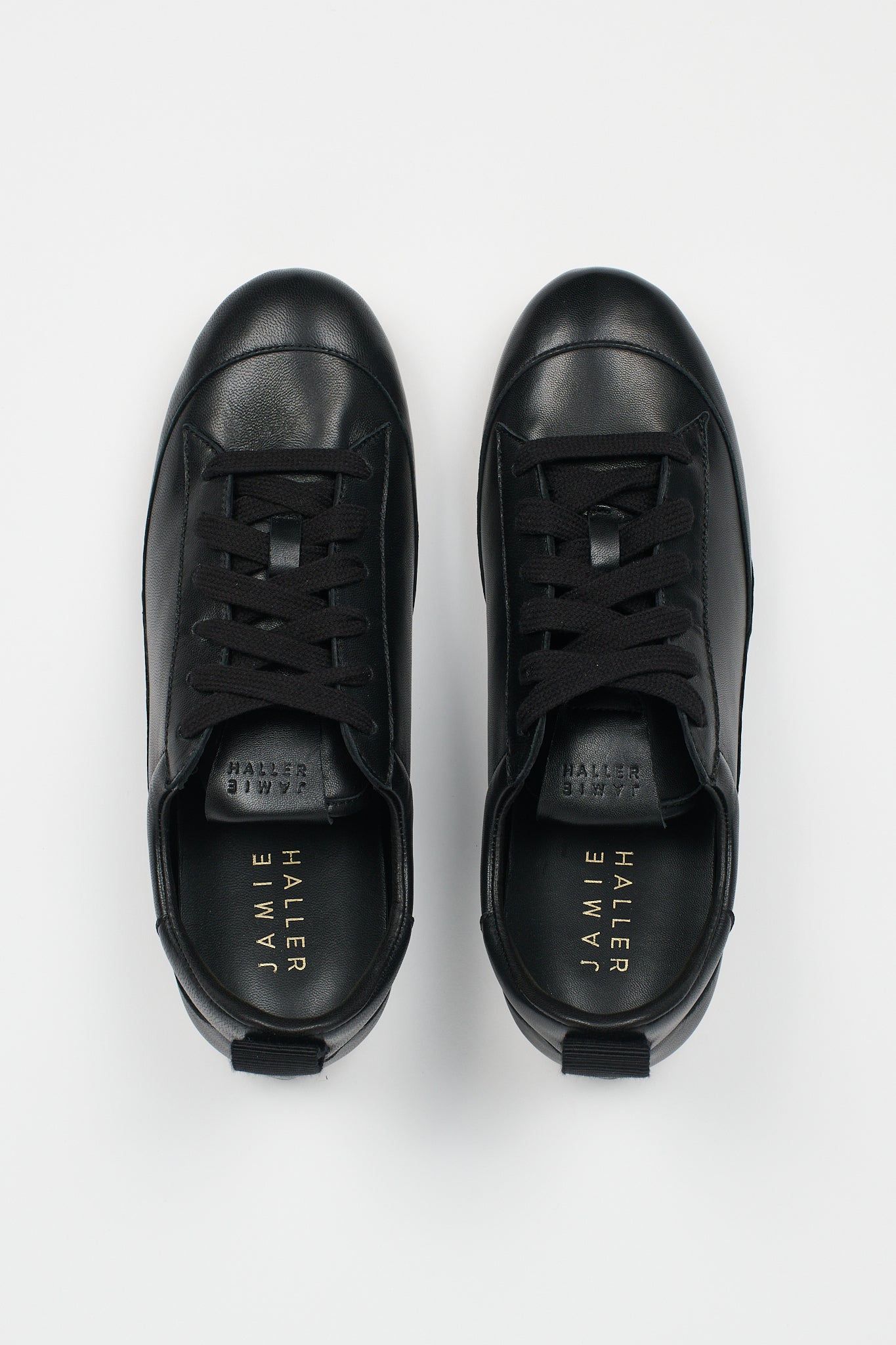A pair of black leather low-top sneakers shown from a top-down view on a light gray background. The brand name 'JAMIE HALLER' is printed in gold on the black insoles.