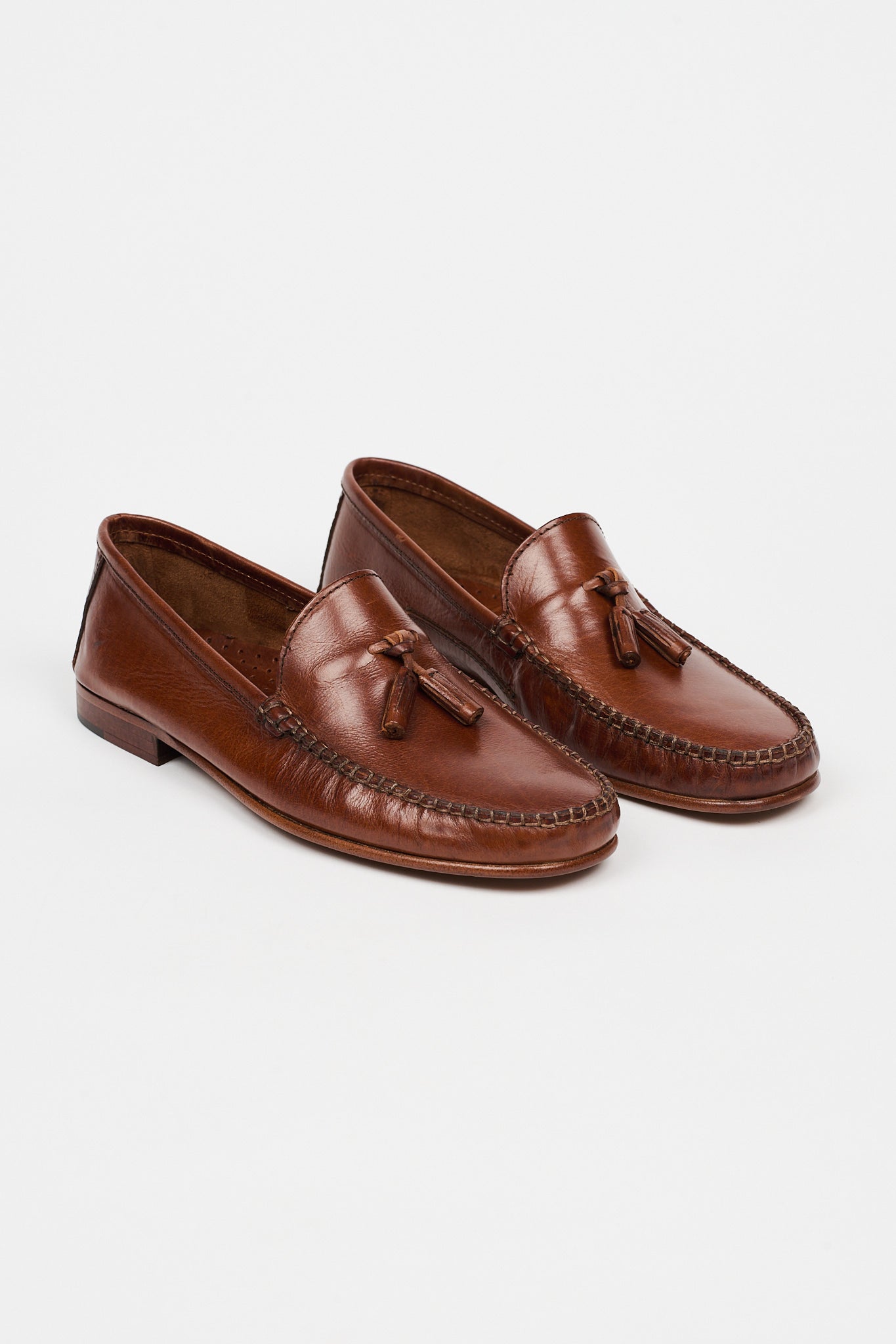 A pair of brown leather moccasin-style loafers with tassel details, photographed on a clean, light gray studio background.