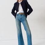 A model stands in a studio wearing a dark navy blazer, a white t-shirt, light-wash blue flare jeans with a brown belt, and dark blue patterned heels.