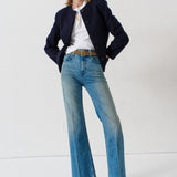 A model stands in a studio wearing a dark navy blazer, a white t-shirt, light-wash blue flare jeans with a brown belt, and dark blue patterned heels.
