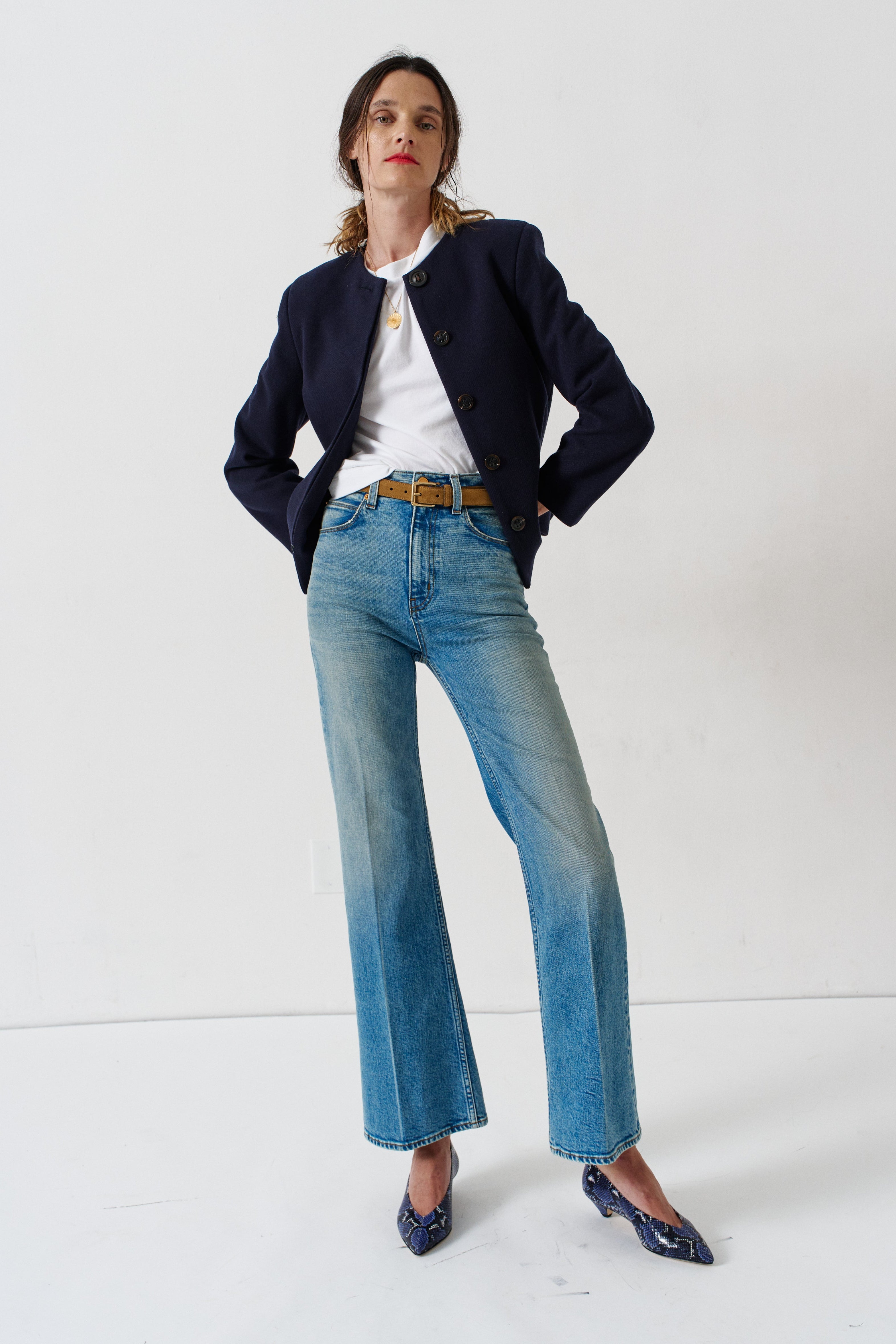 A model stands in a studio wearing a dark navy blazer, a white t-shirt, light-wash blue flare jeans with a brown belt, and dark blue patterned heels.
