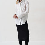 model in a white blouse, black skirt, and black heels against a white wall
