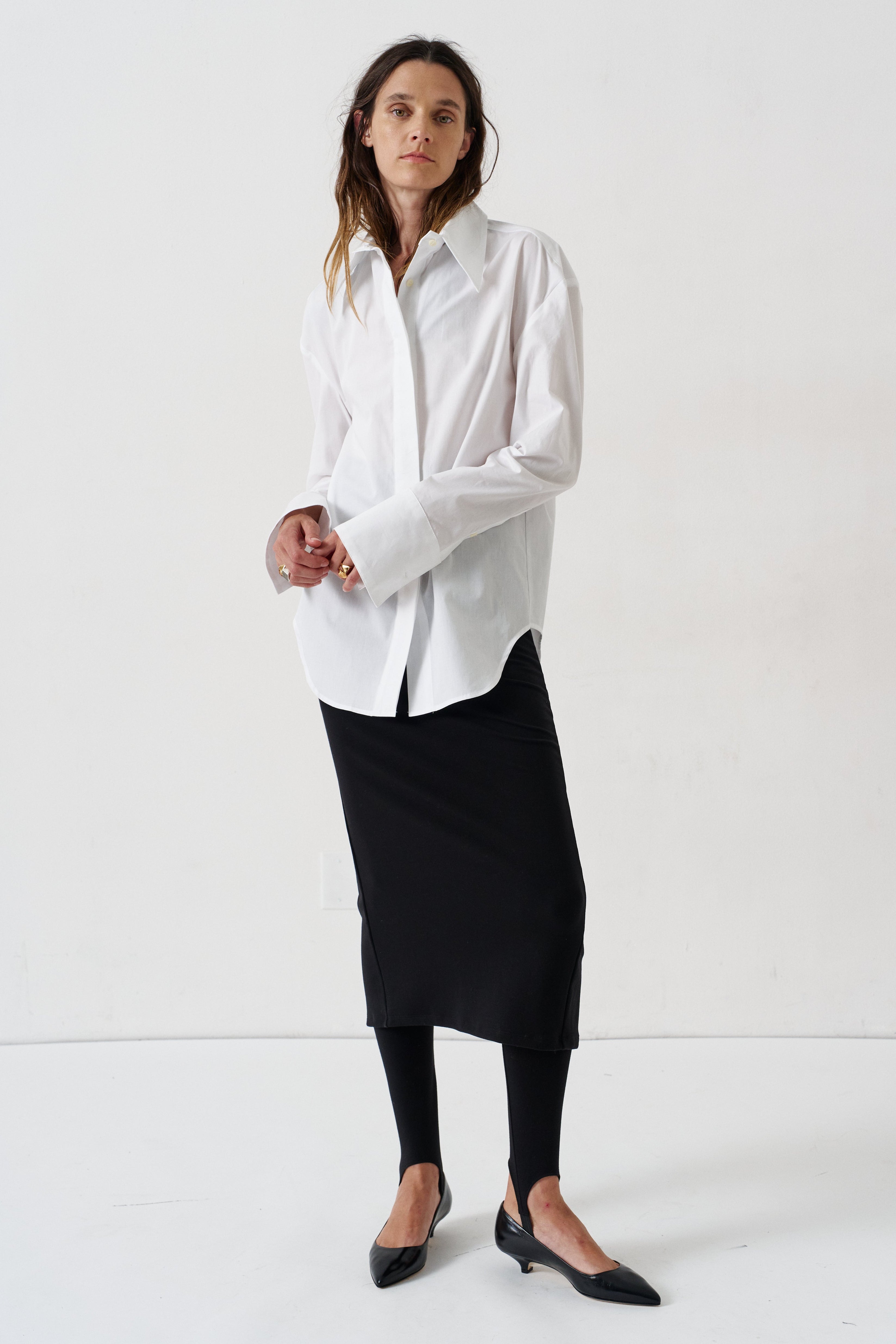 model in a white blouse, black skirt, and black heels against a white wall