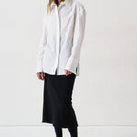 A woman modeling a white long-sleeve button-up shirt with a relaxed, oversized fit, paired with a black midi skirt, black tights, and black pointed flats, set against a plain white studio background.