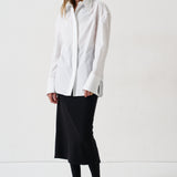 A woman modeling a white long-sleeve button-up shirt with a relaxed, oversized fit, paired with a black midi skirt, black tights, and black pointed flats, set against a plain white studio background.