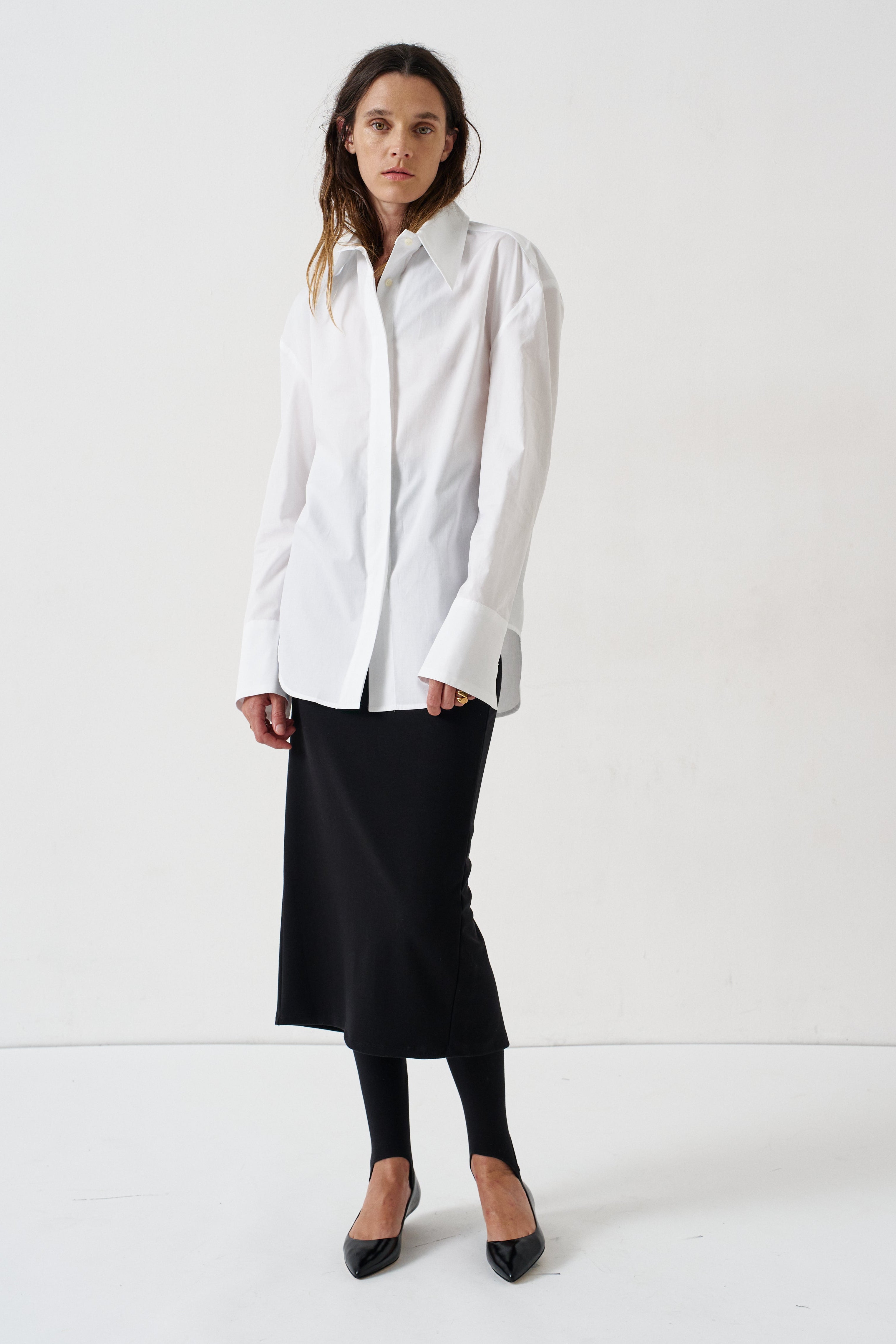 A woman modeling a white long-sleeve button-up shirt with a relaxed, oversized fit, paired with a black midi skirt, black tights, and black pointed flats, set against a plain white studio background.