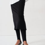 A person wearing a black midi skirt, black tights, and black pointed-toe flats, cropped from the waist down against a plain white studio background.