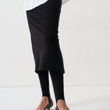A person wearing a black midi skirt, black tights, and black pointed-toe flats, cropped from the waist down against a plain white studio background.