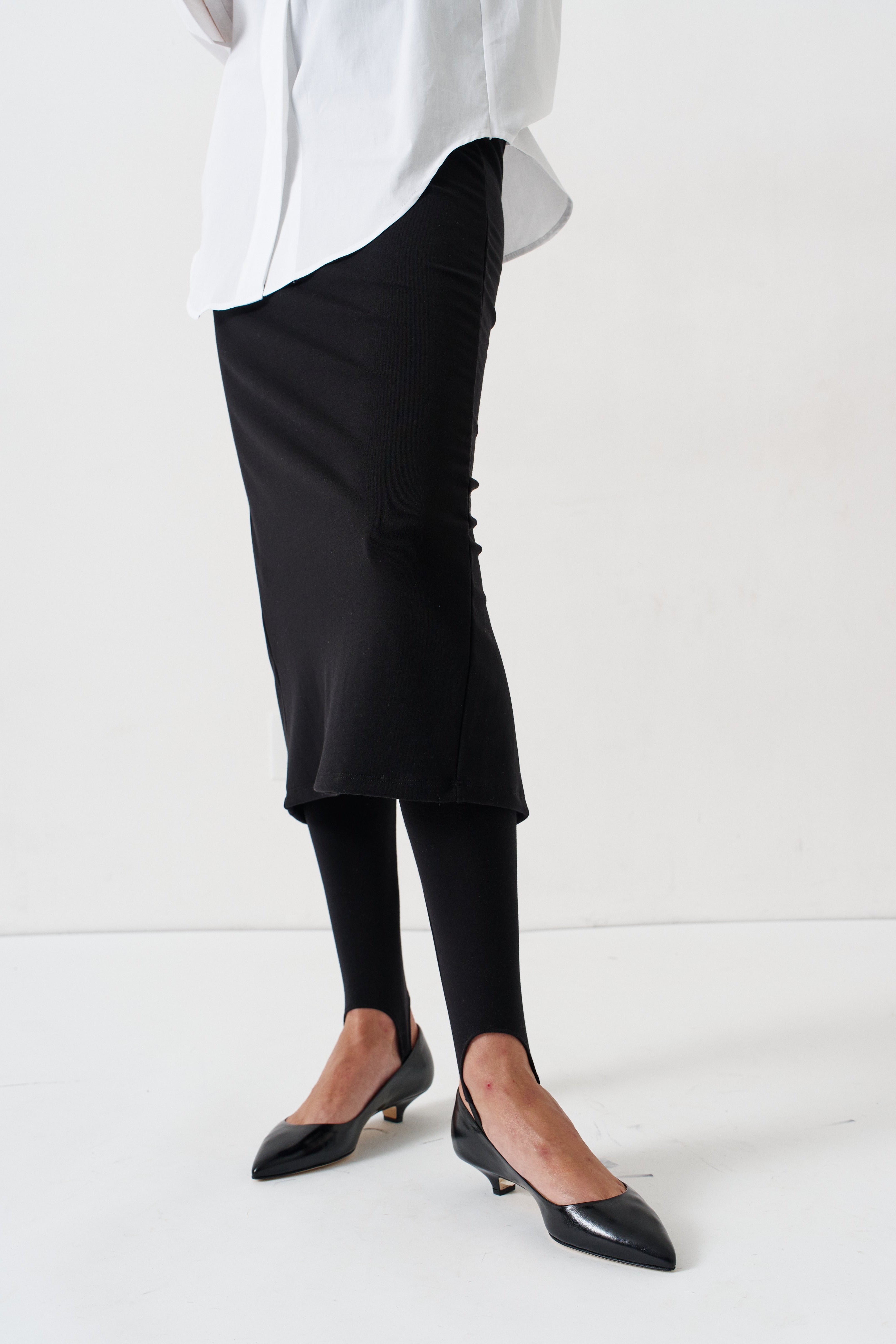 A person wearing a black midi skirt, black tights, and black pointed-toe flats, cropped from the waist down against a plain white studio background.