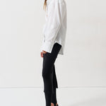 A woman modeling a white long-sleeve button-up shirt with black trousers and black pointed-toe heels, standing in profile against a clean white studio background.