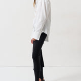 A woman modeling a white long-sleeve button-up shirt with black trousers and black pointed-toe heels, standing in profile against a clean white studio background.