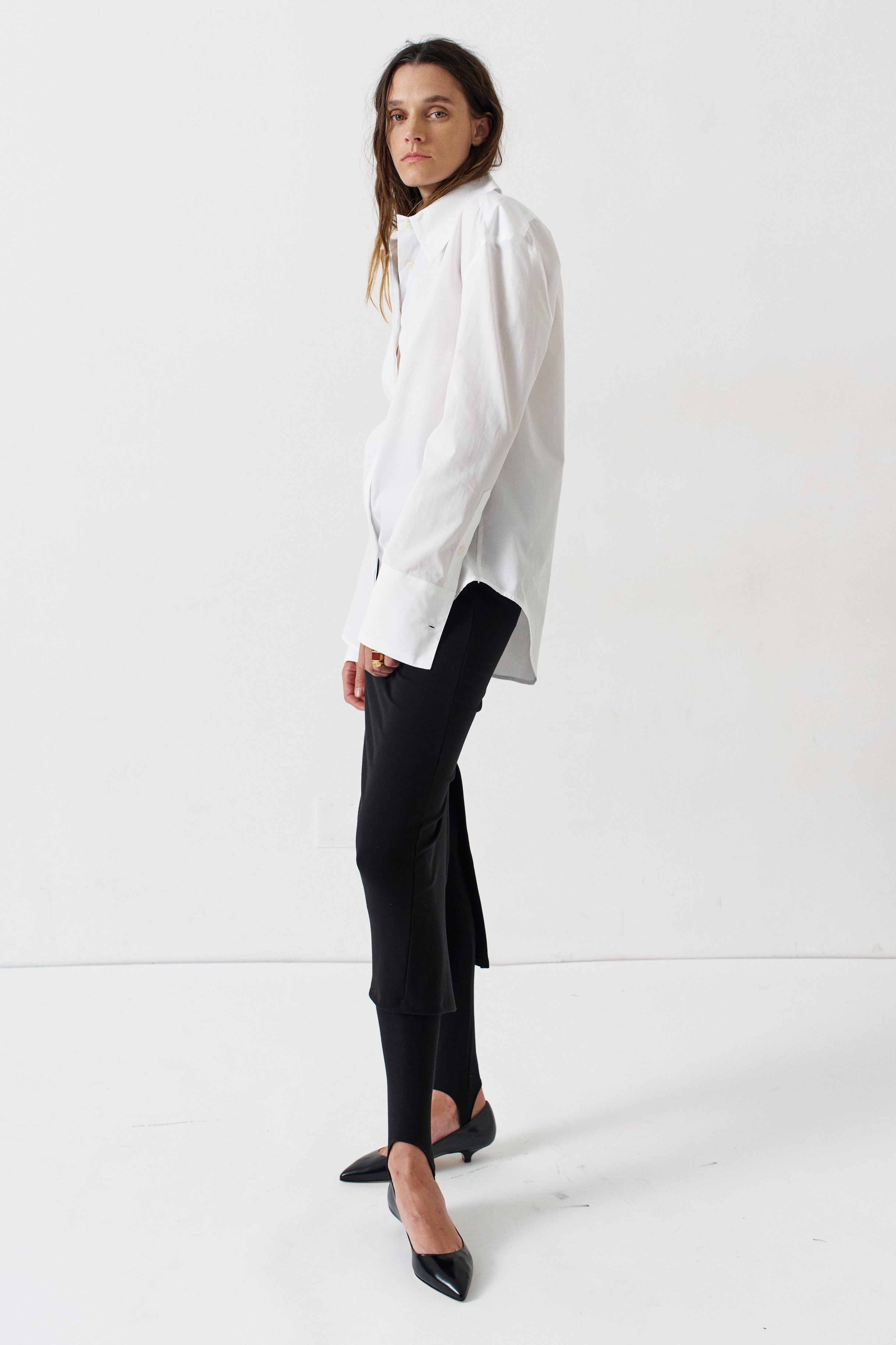 A woman modeling a white long-sleeve button-up shirt with black trousers and black pointed-toe heels, standing in profile against a clean white studio background.