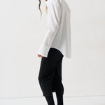 A woman modeling a white oversized button-down shirt paired with black cropped pants, black tights, and black pointed flats, photographed in a bright white studio setting.