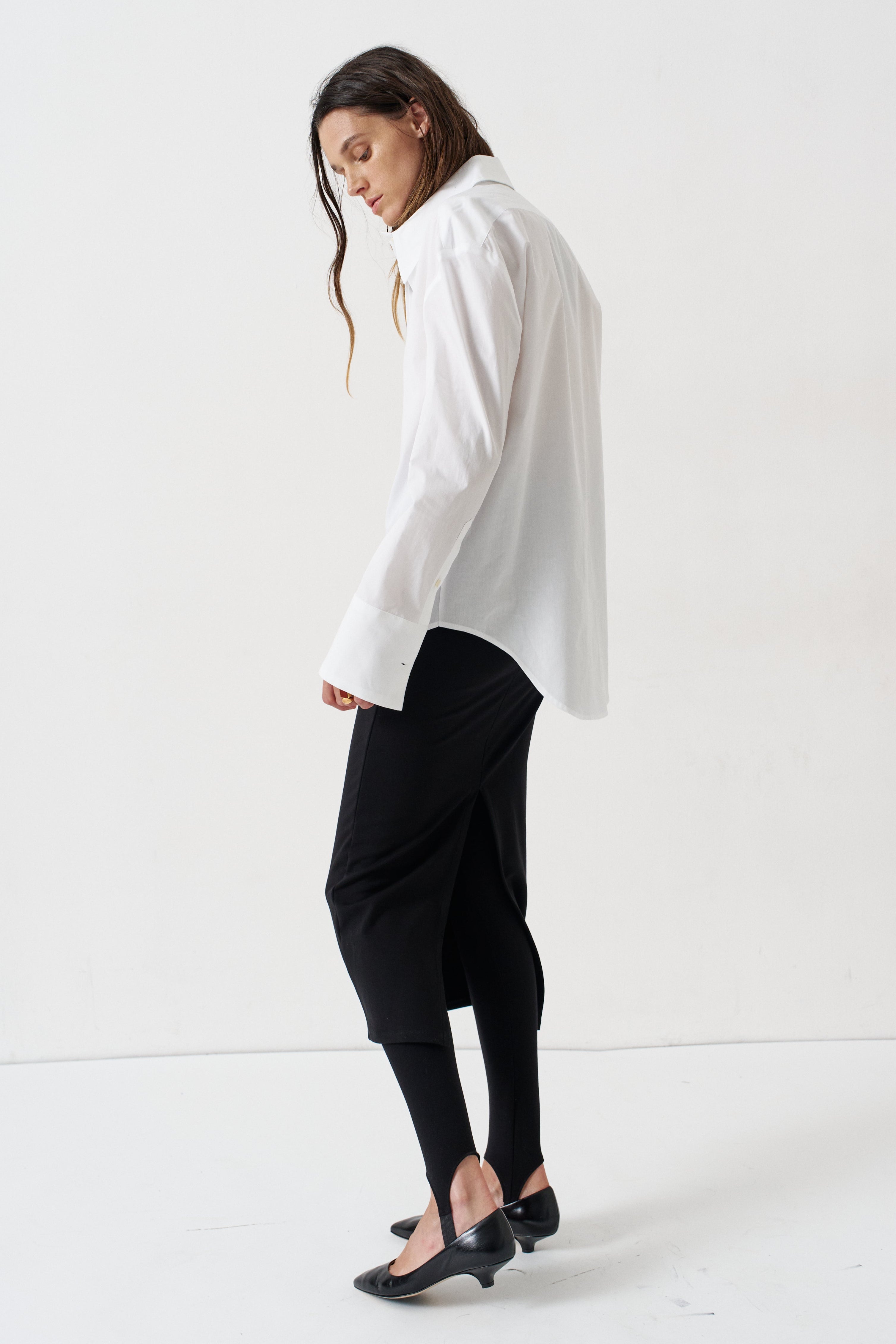 A woman modeling a white oversized button-down shirt paired with black cropped pants, black tights, and black pointed flats, photographed in a bright white studio setting.