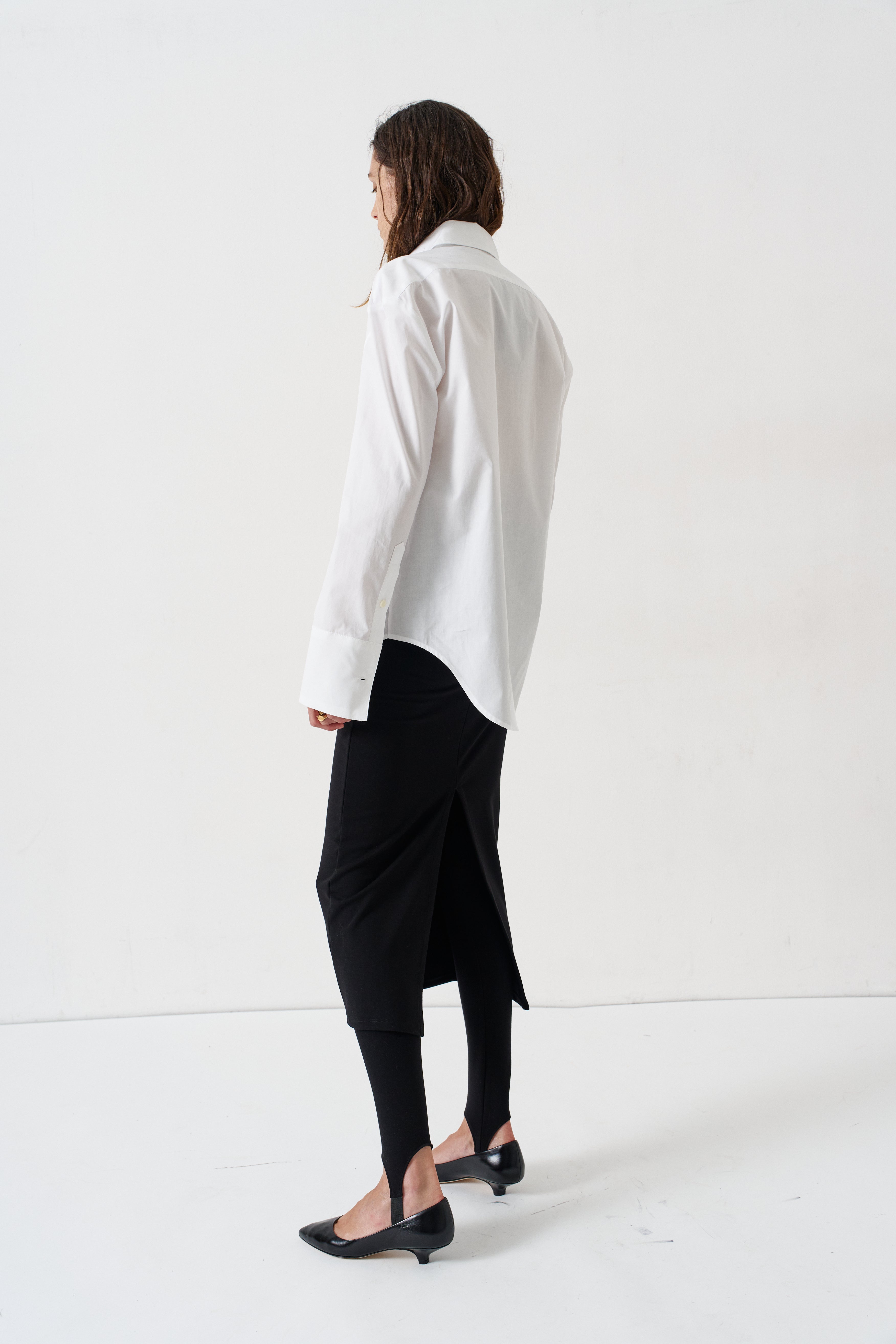 A woman shown from the back wearing a white long-sleeve button-up shirt, a black wrap-style skirt, black tights, and black pointed flats, standing against a plain white studio background.