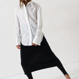A woman modeling a white long-sleeve button-up shirt and a black midi skirt, paired with black pointed-toe heels, against a clean white studio background.