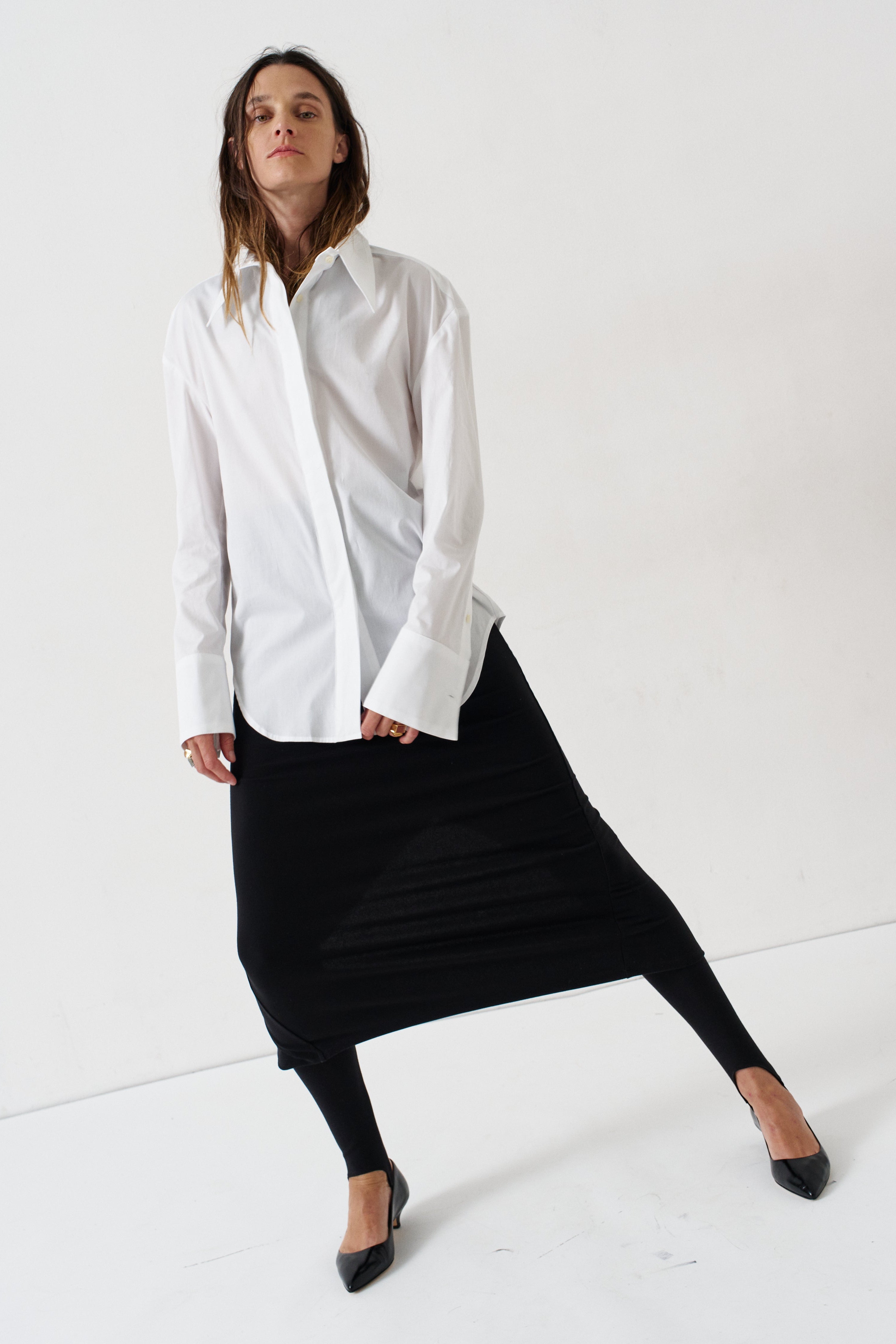 A woman modeling a white long-sleeve button-up shirt and a black midi skirt, paired with black pointed-toe heels, against a clean white studio background.