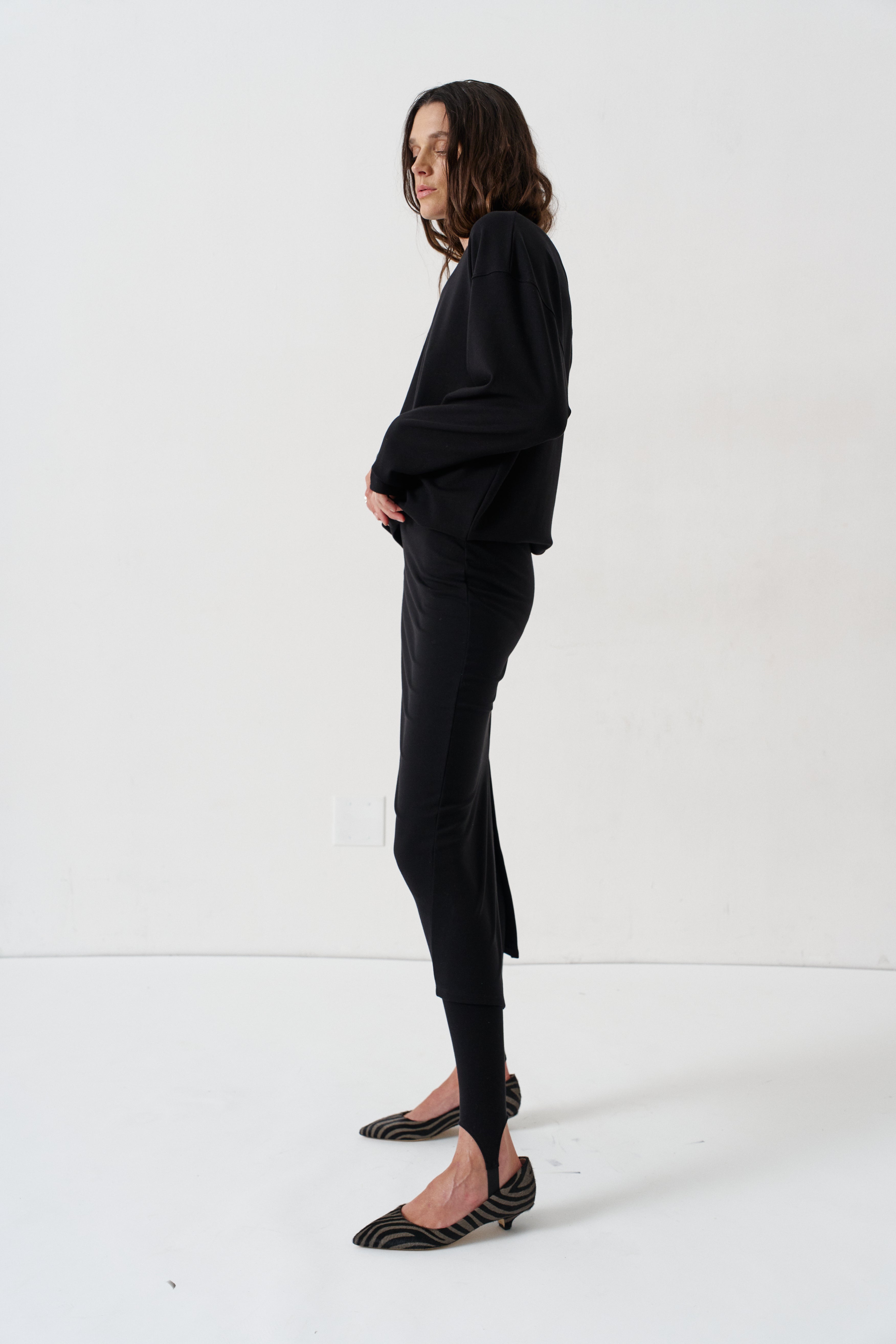 A model is shown in profile wearing a black long-sleeve top, black pants, and black-and-white zebra-patterned pointed-toe flats against a plain white studio background.