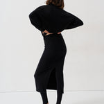 A woman wearing a black long-sleeve midi dress with a side slit, styled with black tights and black-and-white striped kitten heels, photographed from behind against a plain white studio background.