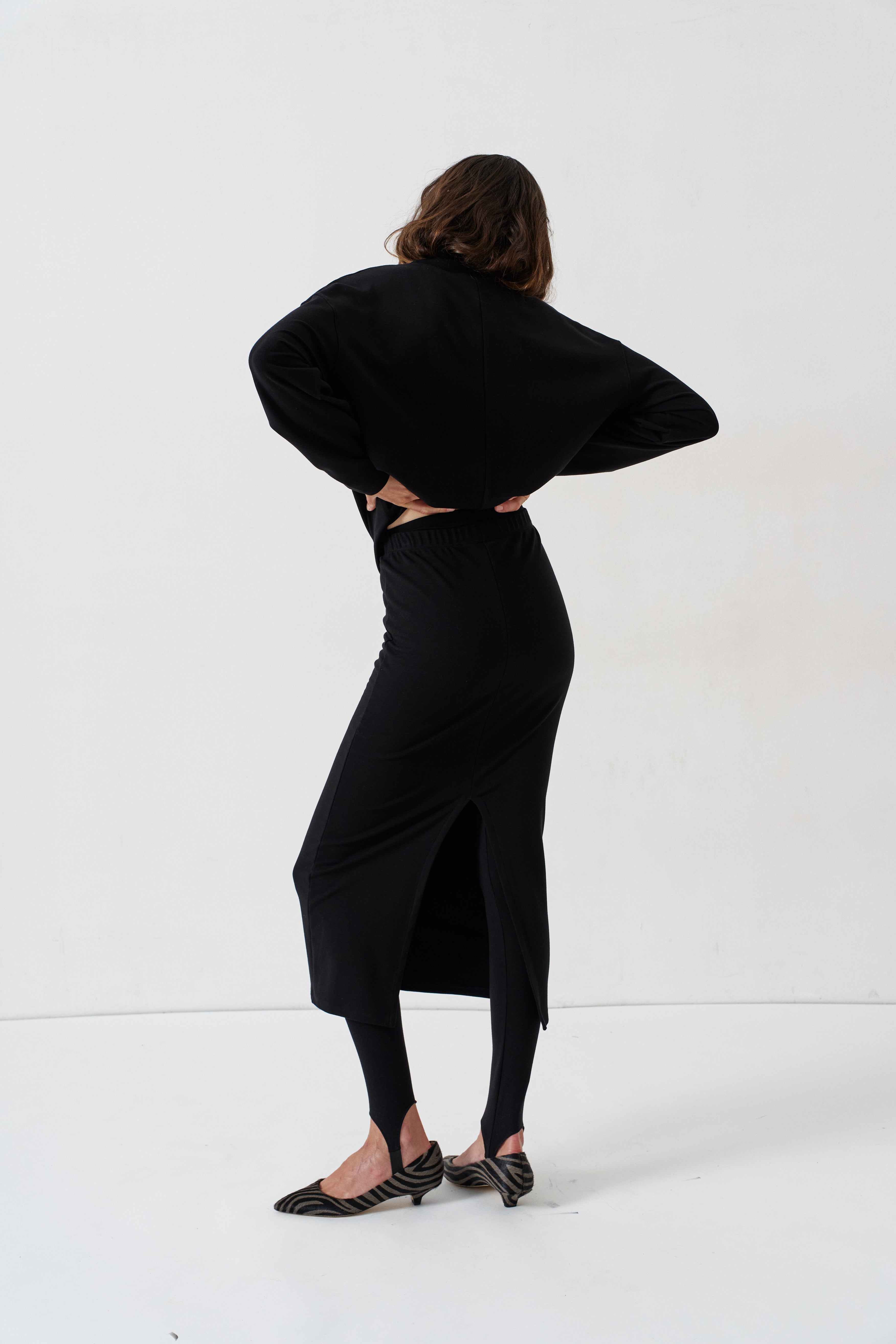 A woman wearing a black long-sleeve midi dress with a side slit, styled with black tights and black-and-white striped kitten heels, photographed from behind against a plain white studio background.