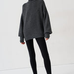 A woman modeling an oversized dark gray turtleneck sweater, paired with black leggings and black-and-white striped kitten heels, against a plain white studio background.